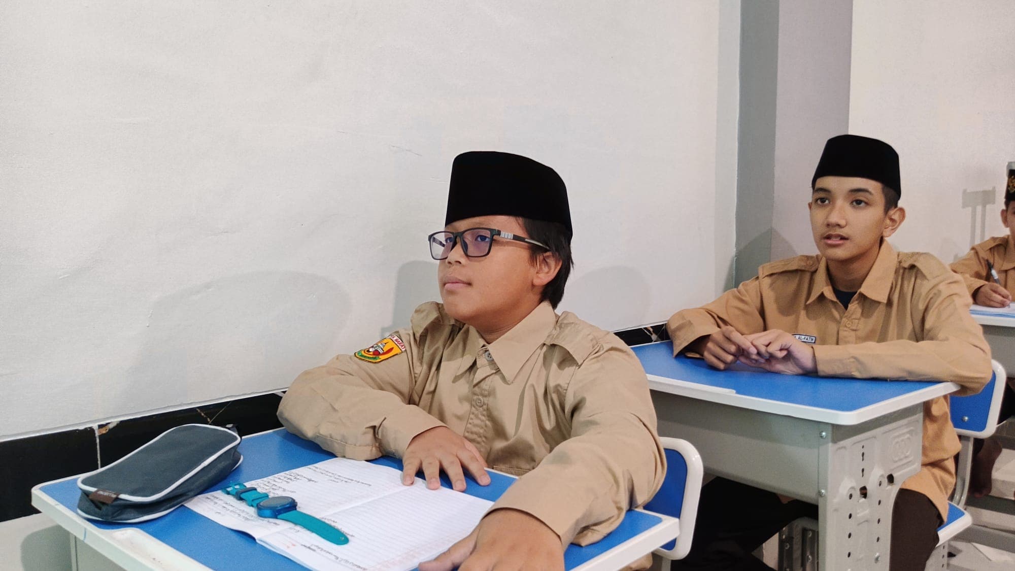 Dua santri MTs dan MA Wadi Mubarak berseragam pramuka menyimak penjelasan ustadz dari bangku kelas