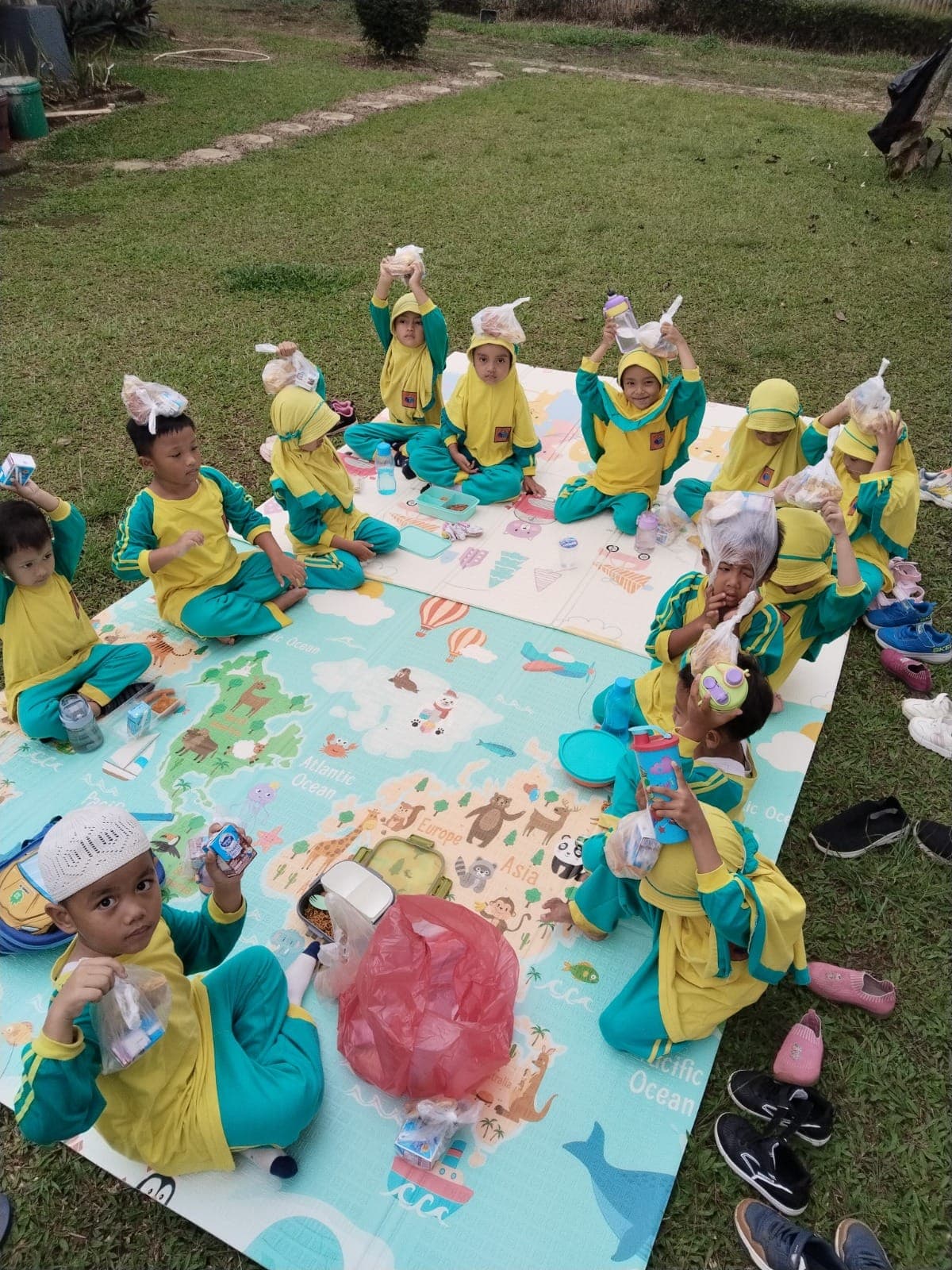 Anak-anak TAUD SAQU duduk di atas tikar bergambar peta di taman sambil menunjukkan bekal mereka