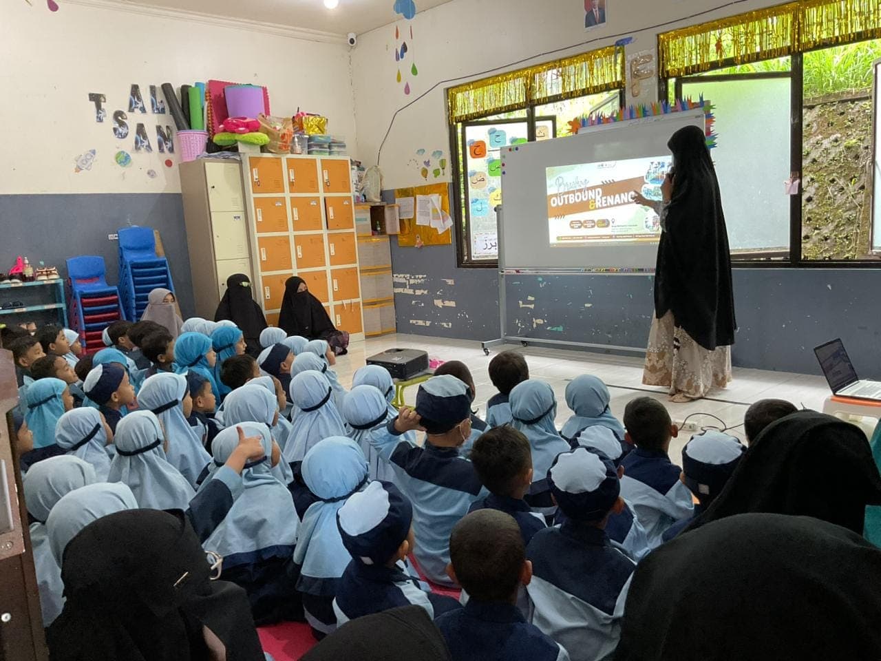 Ustadzah TAUD SAQU menerangkan materi lewat layar di depan anak-anak yang duduk rapi di lantai kelas